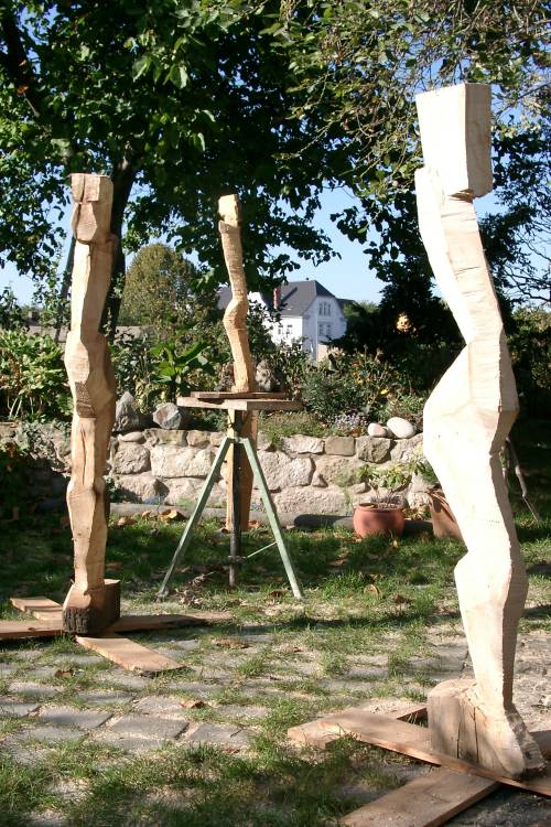 Drei Holzskulpturen im Garten. Zwei große im Vordergrund, im Hintergrund eine kleinere auf einem Sockel.